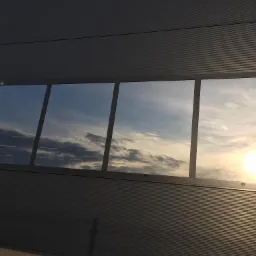 Cztery okna w szarej, metalowej ścianie budynku przemysłowego odbijające błękitne niebo z chmurami i zachodzące słońce; widoczna kamera przemysłowa w rogu.