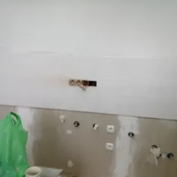 Fragment ściany z białymi płytkami, widoczne przygotowane otwory pod gniazdka elektryczne i puszka instalacyjna z wystającymi przewodami, żółta poziomica przy ścianie, oraz worki foliowe i kubek...