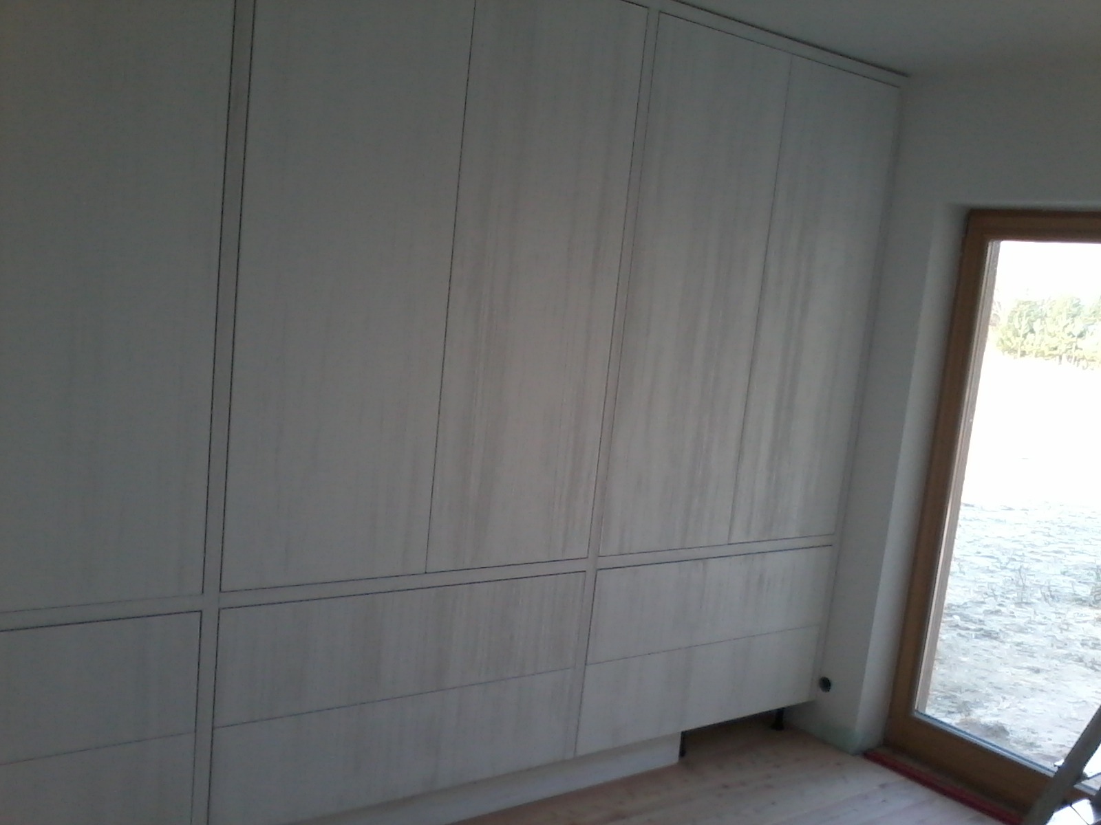 Duża, jasna szafa wnękowa z drewnianymi frontami, widoczna z boku, z oknem w tle.