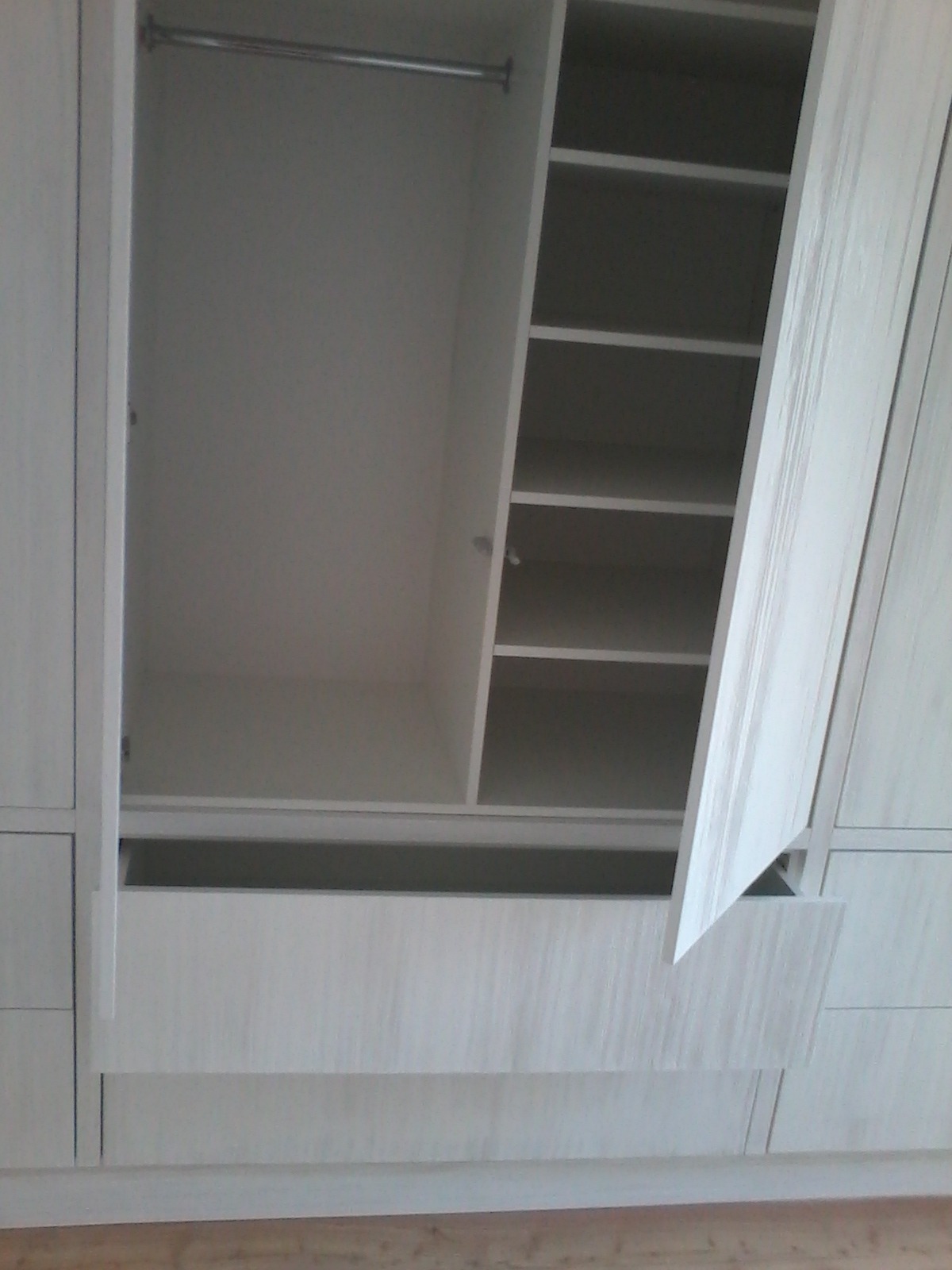 Wnętrze jasnej, drewnopodobnej szafy z drzwiami uchylonymi, widoczne półki i drążek na ubrania, szuflada częściowo wysunięta.