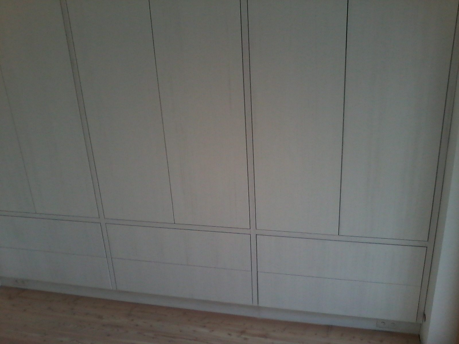 Biała szafa wnękowa z szufladami i drzwiami, minimalistyczny design, widoczne listwy przypodłogowe i fragment podłogi z jasnego drewna.