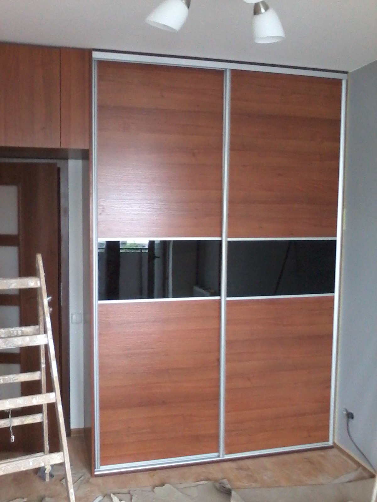 Dwu-drzwiowa szafa przesuwna z drewnopodobnymi panelami i czarnymi, szklanymi wstawkami, widoczna drabina malarska w tle, sugerująca montaż w trakcie realizacji.