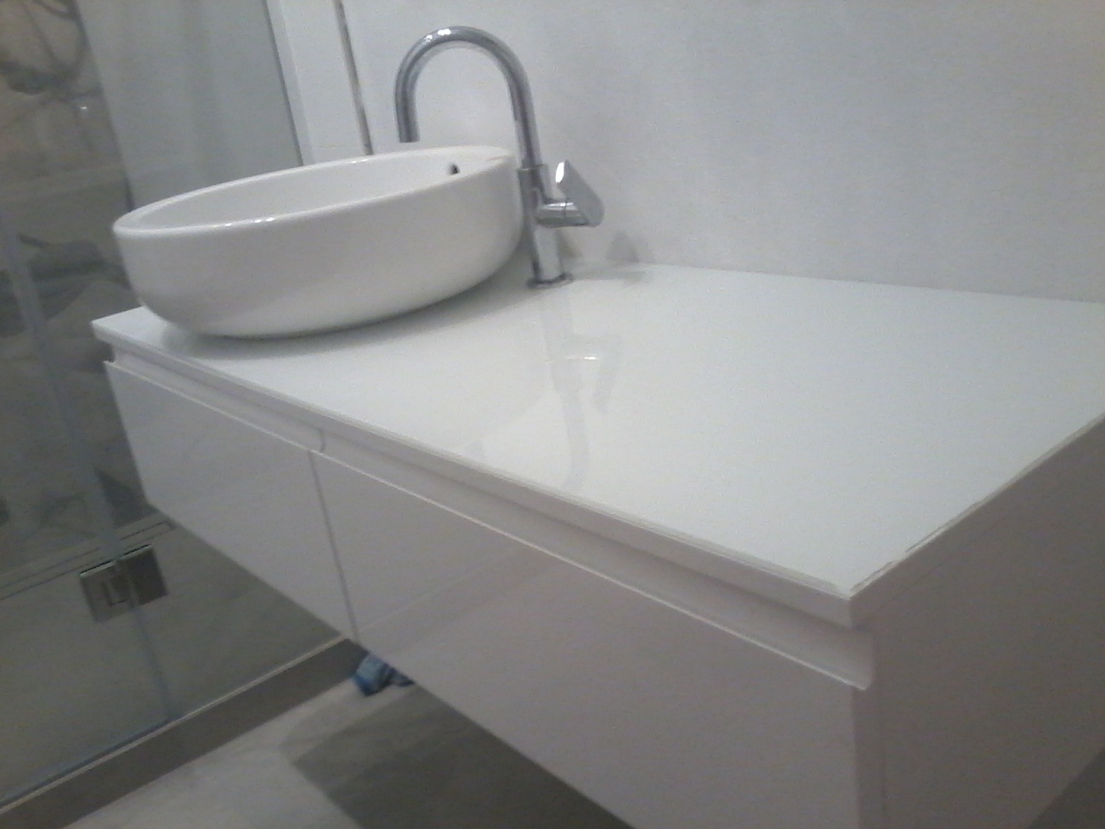 Biała umywalka nablatowa o owalnym kształcie z chromowaną baterią, zamontowana na białej szafce pod umywalkę z gładkimi frontami, widoczny fragment kabiny prysznicowej w tle.