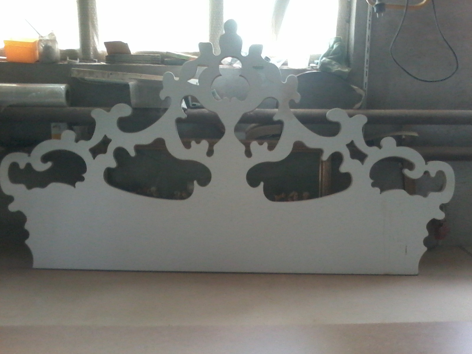 Biały, ozdobny zagłówek łóżka z wyciętymi wzorami, w warsztacie, przed oknem i grzejnikiem.