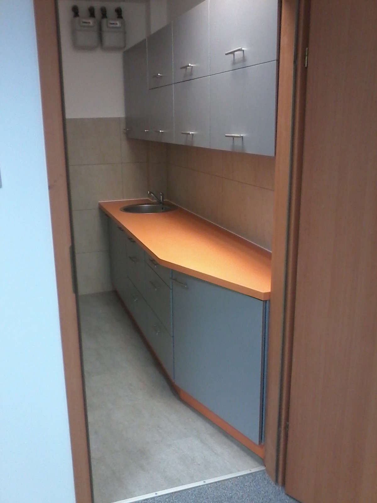 Szare szafki kuchenne z pomarańczowym blatem i metalowym zlewozmywakiem, widoczne zza framugi drzwi.