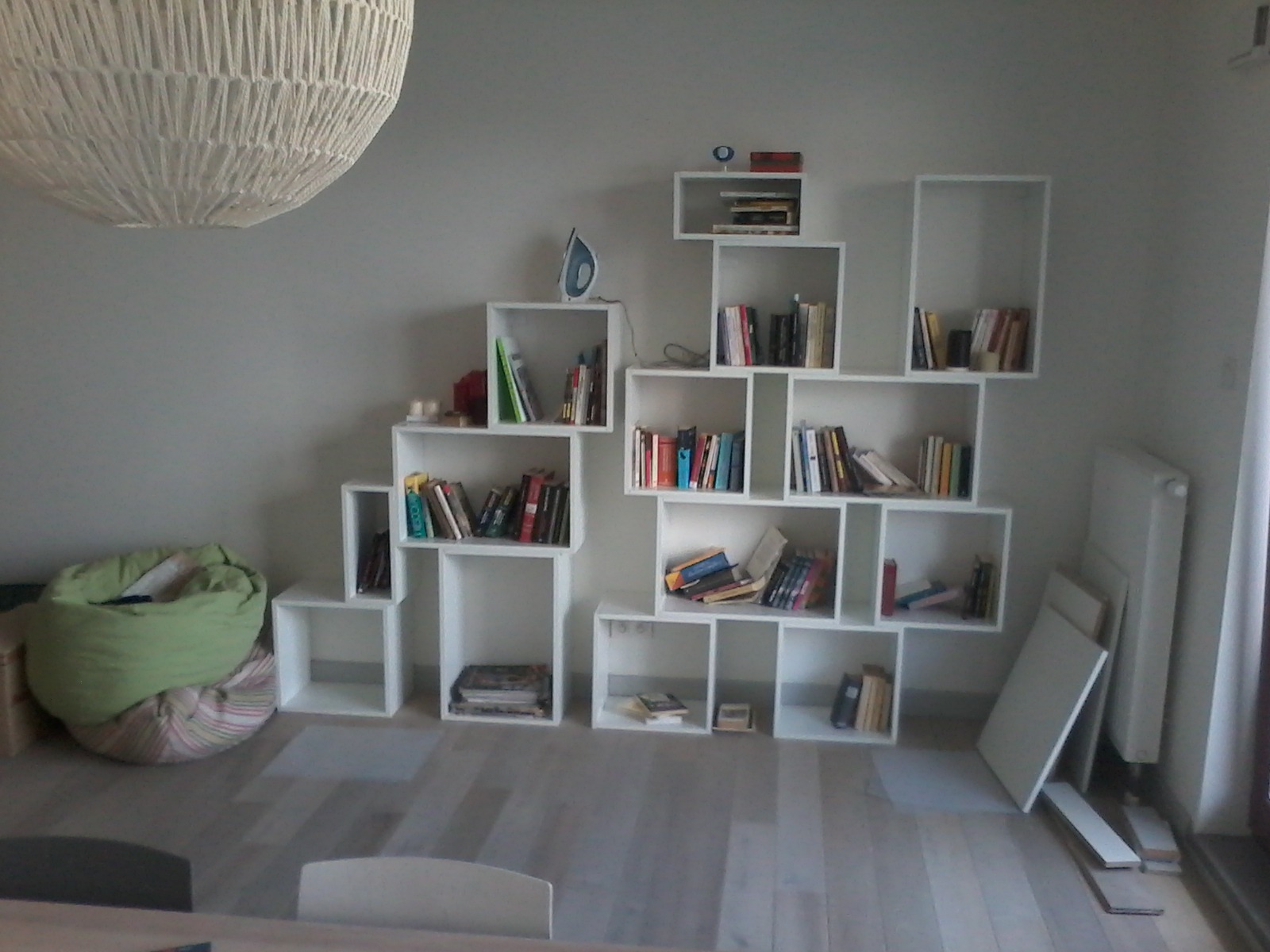 Biała, modułowa półka na książki, ułożona asymetrycznie, wypełniona książkami i ozdobami, obok pufa w paski i lampa wisząca z grubego sznurka