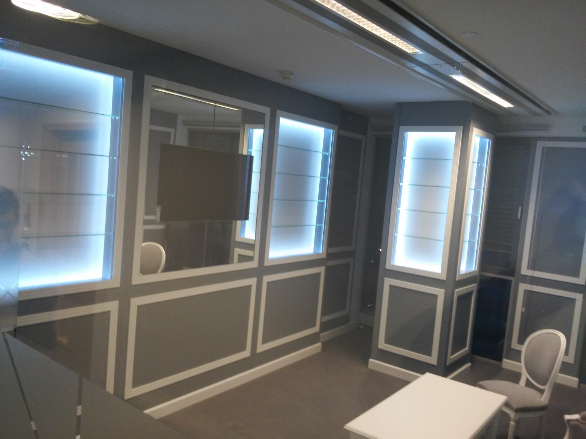 Szare ściany z wbudowanymi podświetlanymi gablotami z szklanymi półkami, lustro, stolik i krzesła w pomieszczeniu.