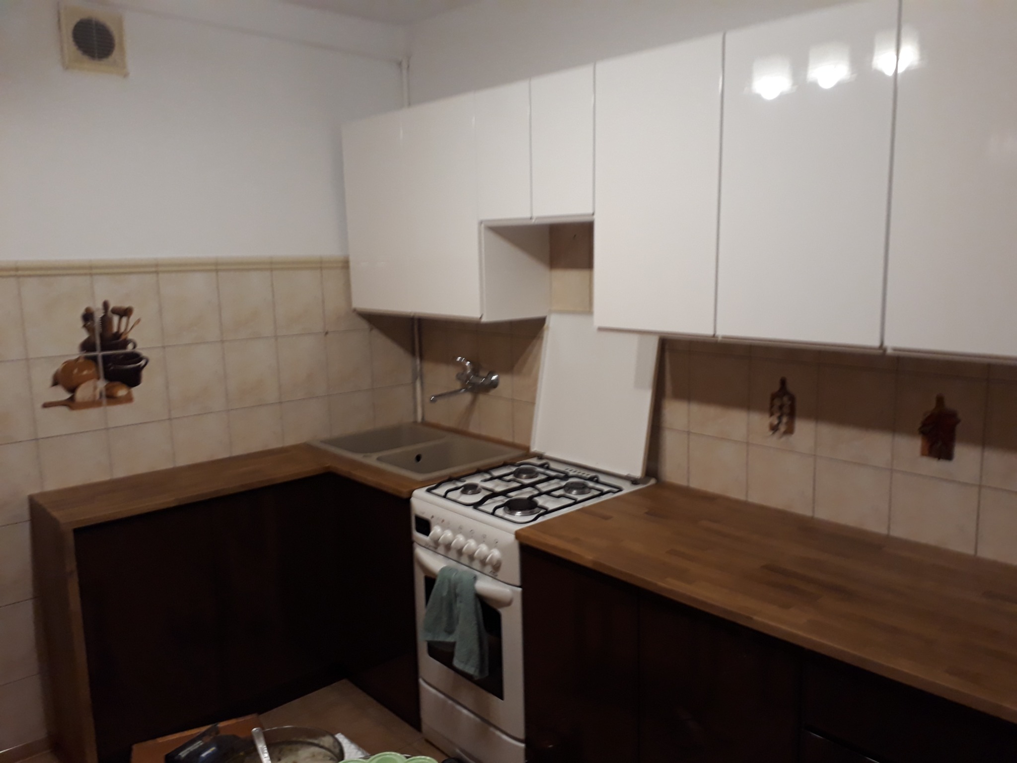 Wnętrze kuchni z białymi szafkami wiszącymi, brązowymi szafkami dolnymi z drewnianym blatem, białą kuchenką gazową z ręcznikiem i beżowymi płytkami na ścianie.