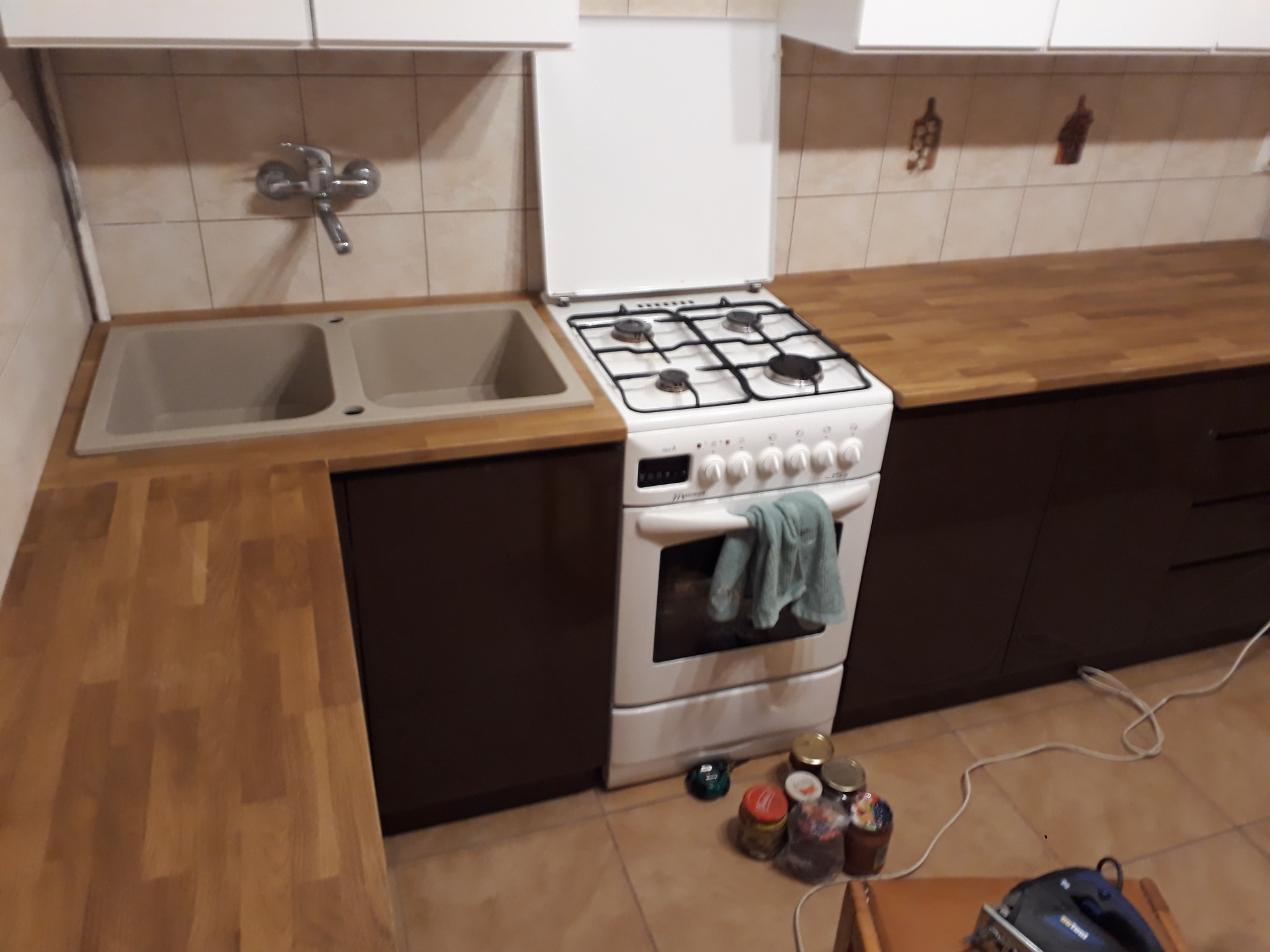 Fragment kuchni z drewnianym blatem, zlewozmywakiem, kuchenką gazową z ręcznikiem i szafkami w odcieniach brązu, widoczne słoiki i elektronarzędzie na podłodze.