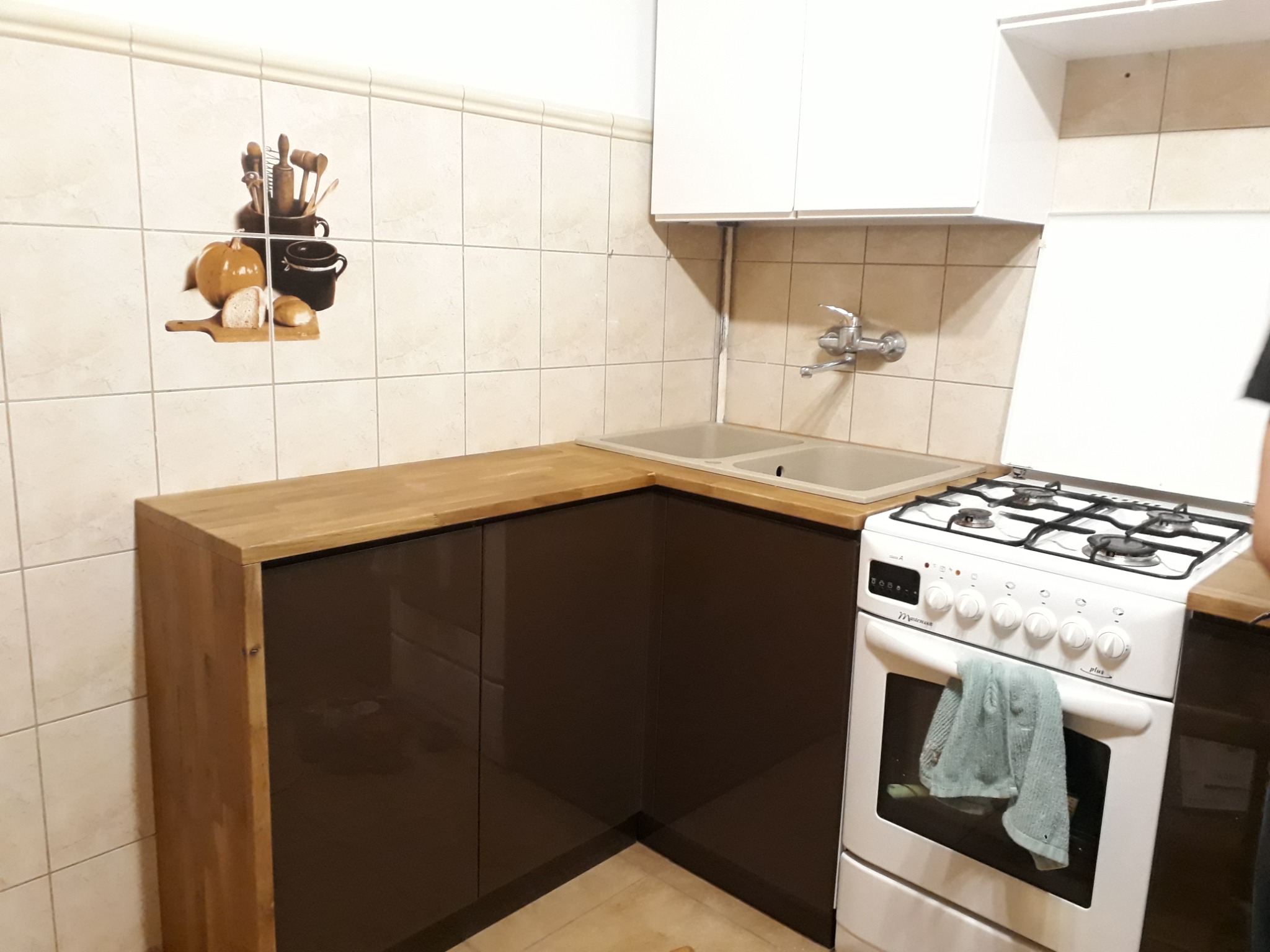 Fragment zabudowy kuchennej z brązowymi, lakierowanymi frontami, drewnianym blatem i białą kuchenką gazową z zawieszoną szmatką. Na ścianie płytki z dekoracyjnym motywem.