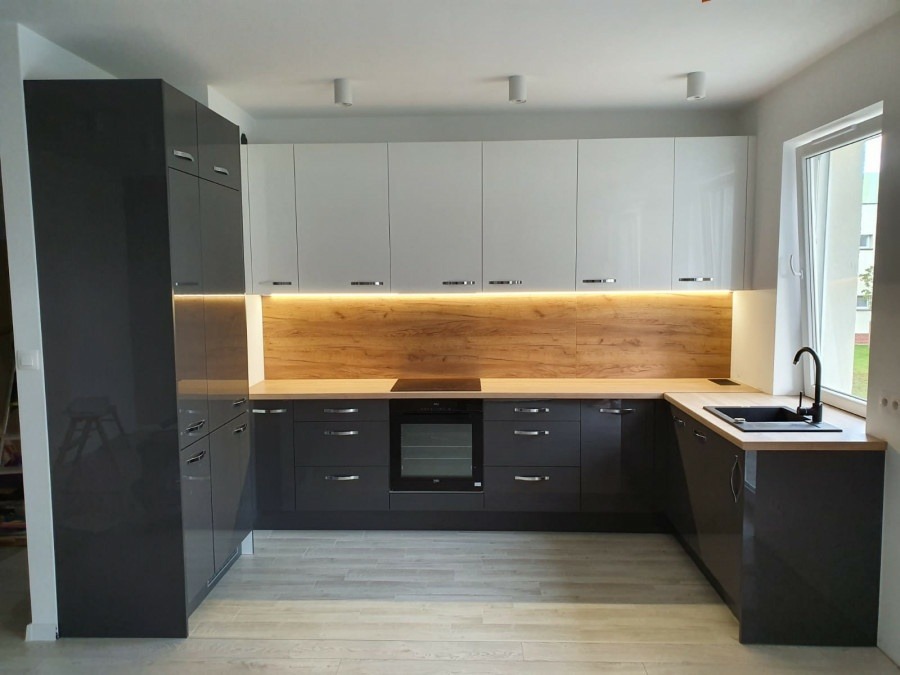 Nowoczesna kuchnia z szarymi dolnymi szafkami i białymi górnymi, drewnianym panelem nad blatem i oświetleniem LED pod górnymi szafkami, z widocznym oknem po prawej stronie.