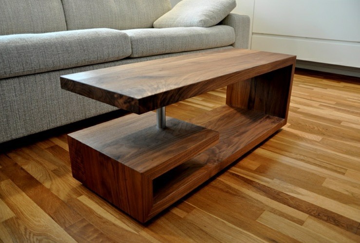 Stolik kawowy z drewna orzechowego o nowoczesnej konstrukcji, z blatem wspartym na metalowej nodze, z dolną półką tworzącą otwartą przestrzeń, na tle parkietu i beżowej sofy.