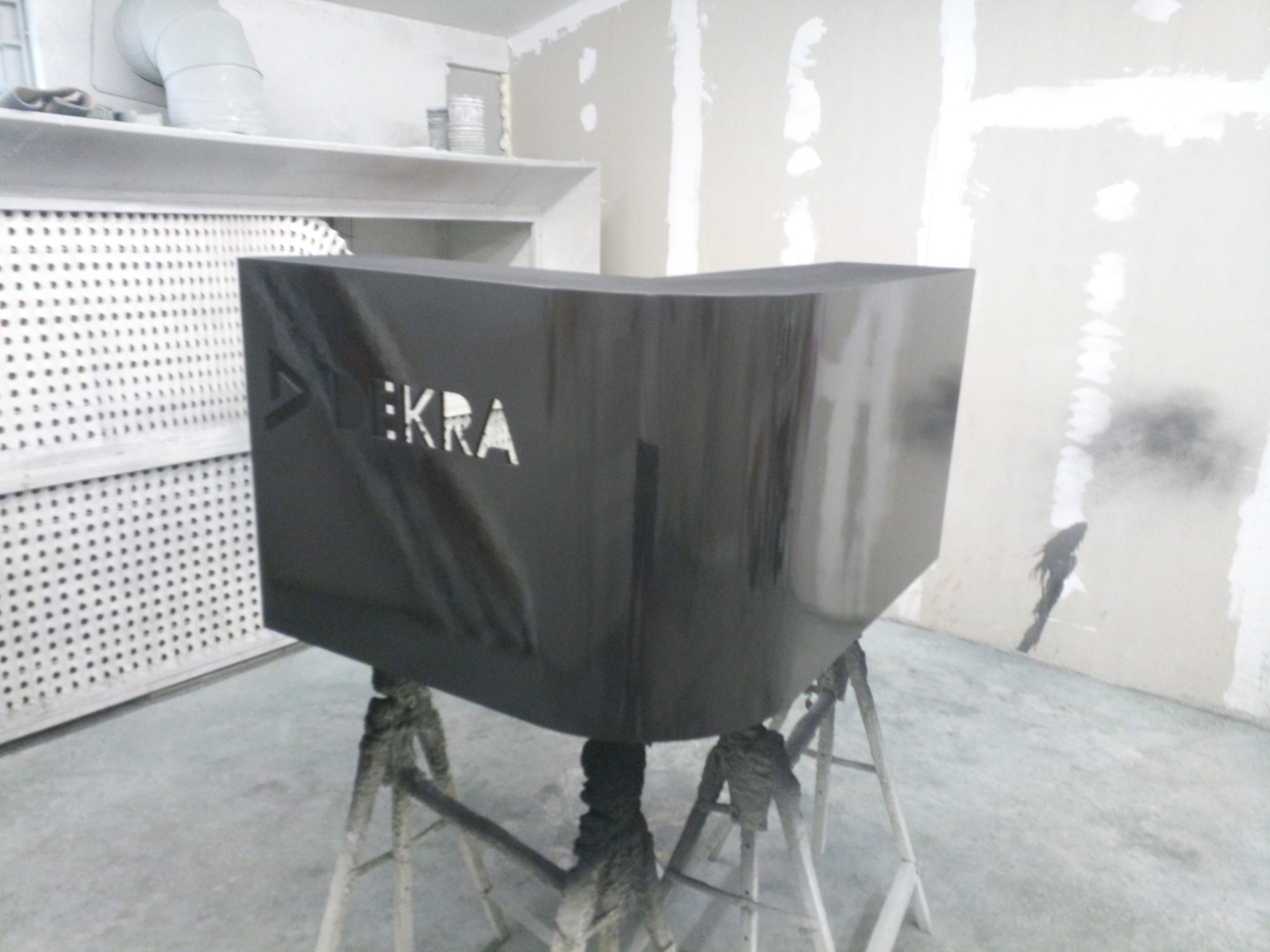 Ciemny, lakierowany blat z wyciętym napisem DEKRA, ustawiony na kozłach w warsztacie, ściany w trakcie malowania.