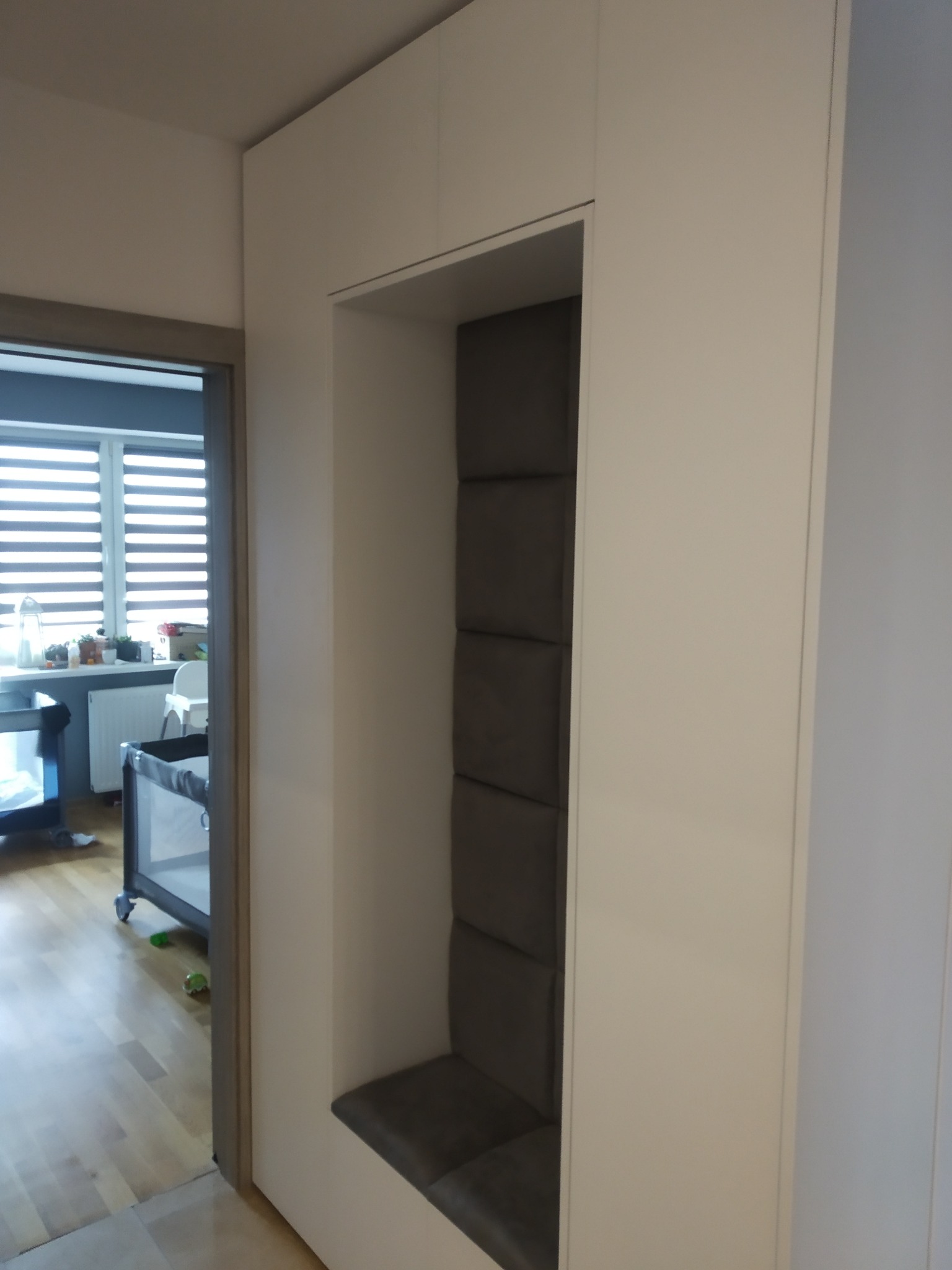 Wnęka w zabudowie meblowej z tapicerowanym siedziskiem i panelem ściennym w kolorze szarym, widok na pokój dziecięcy z łóżeczkiem.