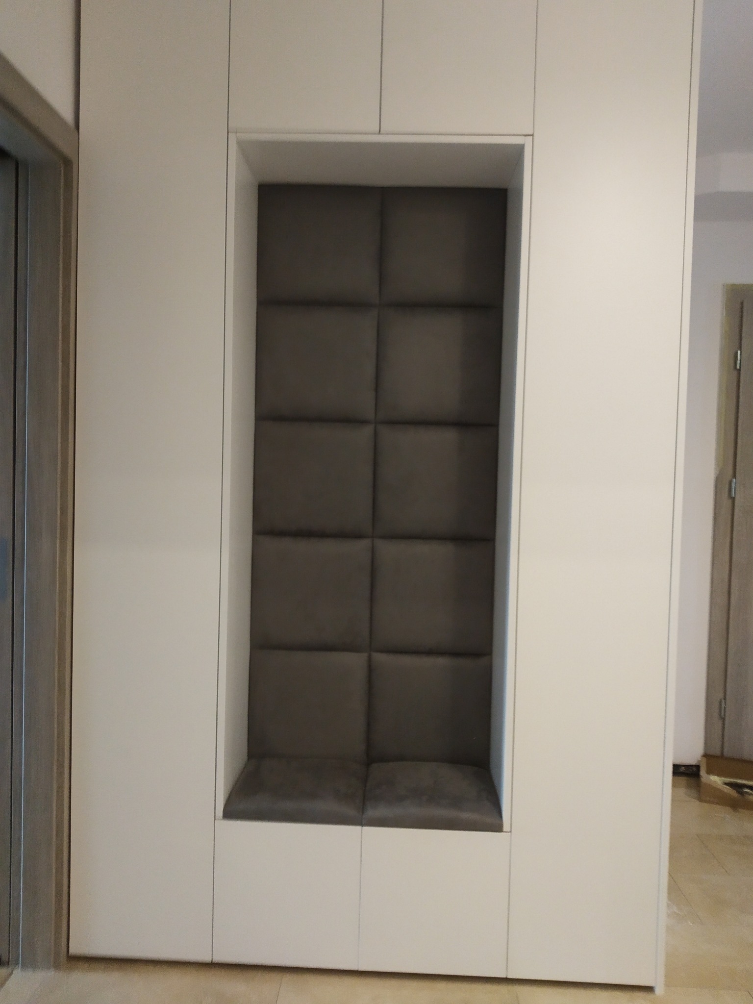 Biała zabudowa meblowa z wnęką wyłożoną szarą tapicerką w kwadraty, tworzącą siedzisko.