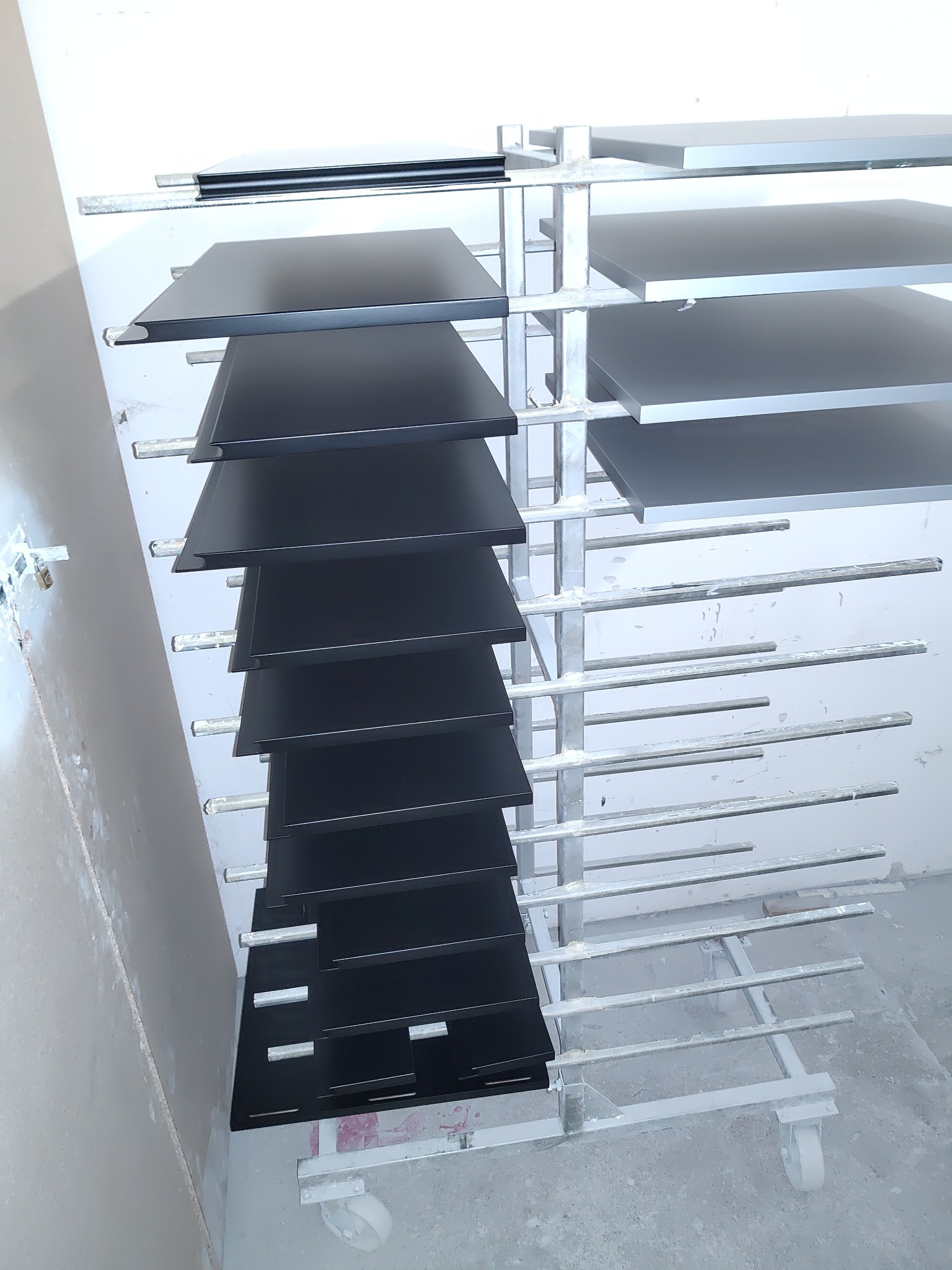 Pomalowane na czarno i szaro płyty, prawdopodobnie blaty lub fronty, suszone na metalowym regale w warsztacie.