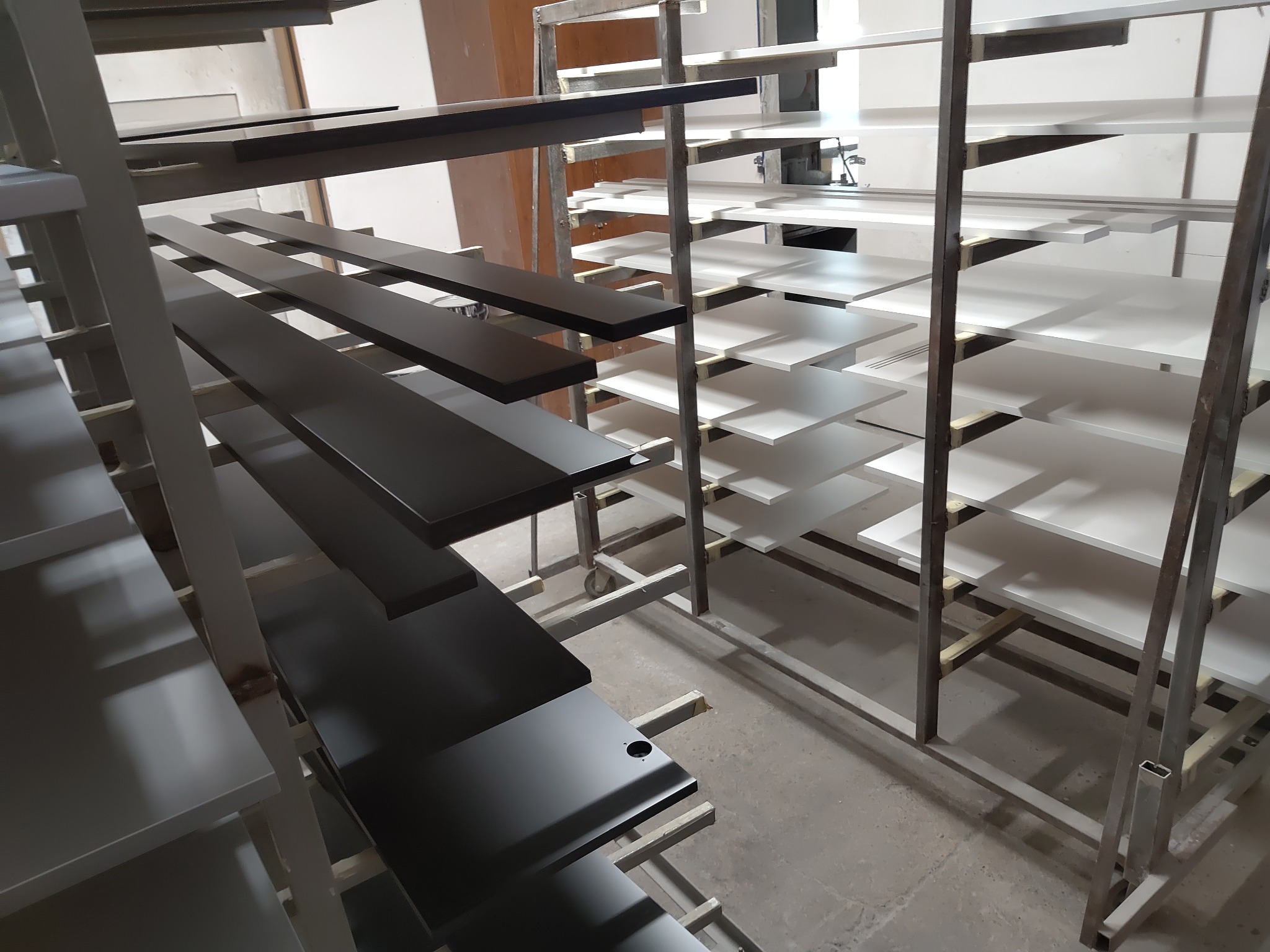 Elementy meblowe, świeżo polakierowane na czarno i biało, schnące na metalowym regale w lakierni.