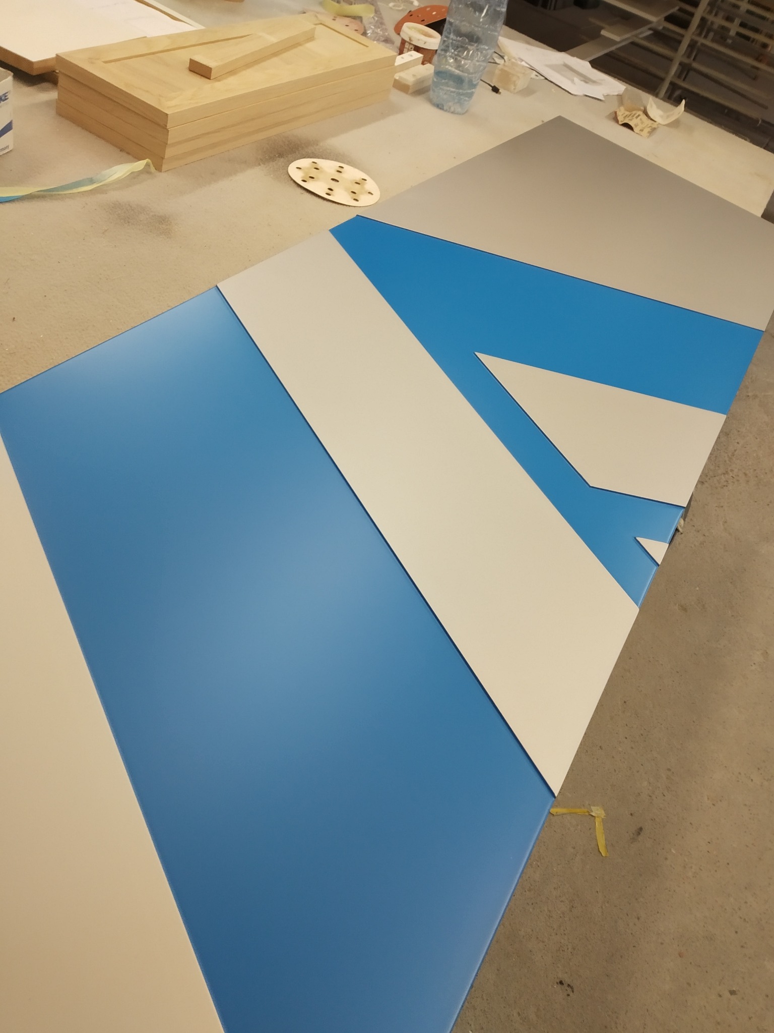 Pomalowany na niebiesko blat z naklejonym wzorem z białych i szarych trójkątów, leżący na podłodze w warsztacie stolarskim, obok leżą elementy drewniane i butelka wody.
