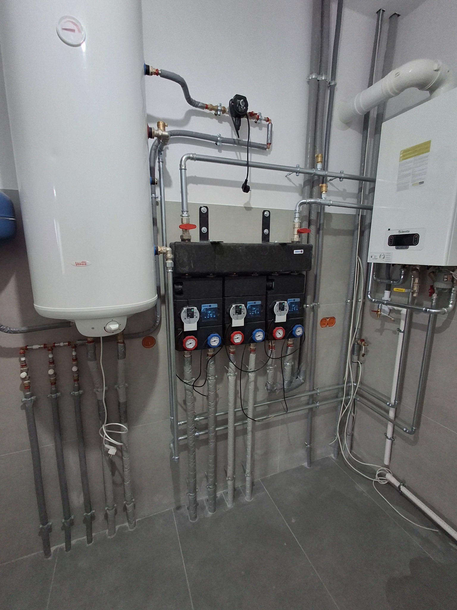 Nowoczesna kotłownia z białym zasobnikiem ciepłej wody, czarnym rozdzielaczem i kotłem gazowym. Szare rury i płytki. Widoczne manometry i zawory.