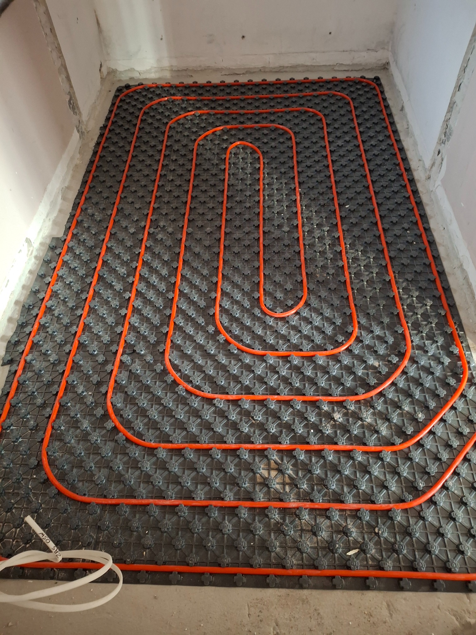 Pomarańczowe rury ogrzewania podłogowego ułożone spiralnie na czarnej macie izolacyjnej w surowym pomieszczeniu. Widoczne fragmenty ścian i przewody.