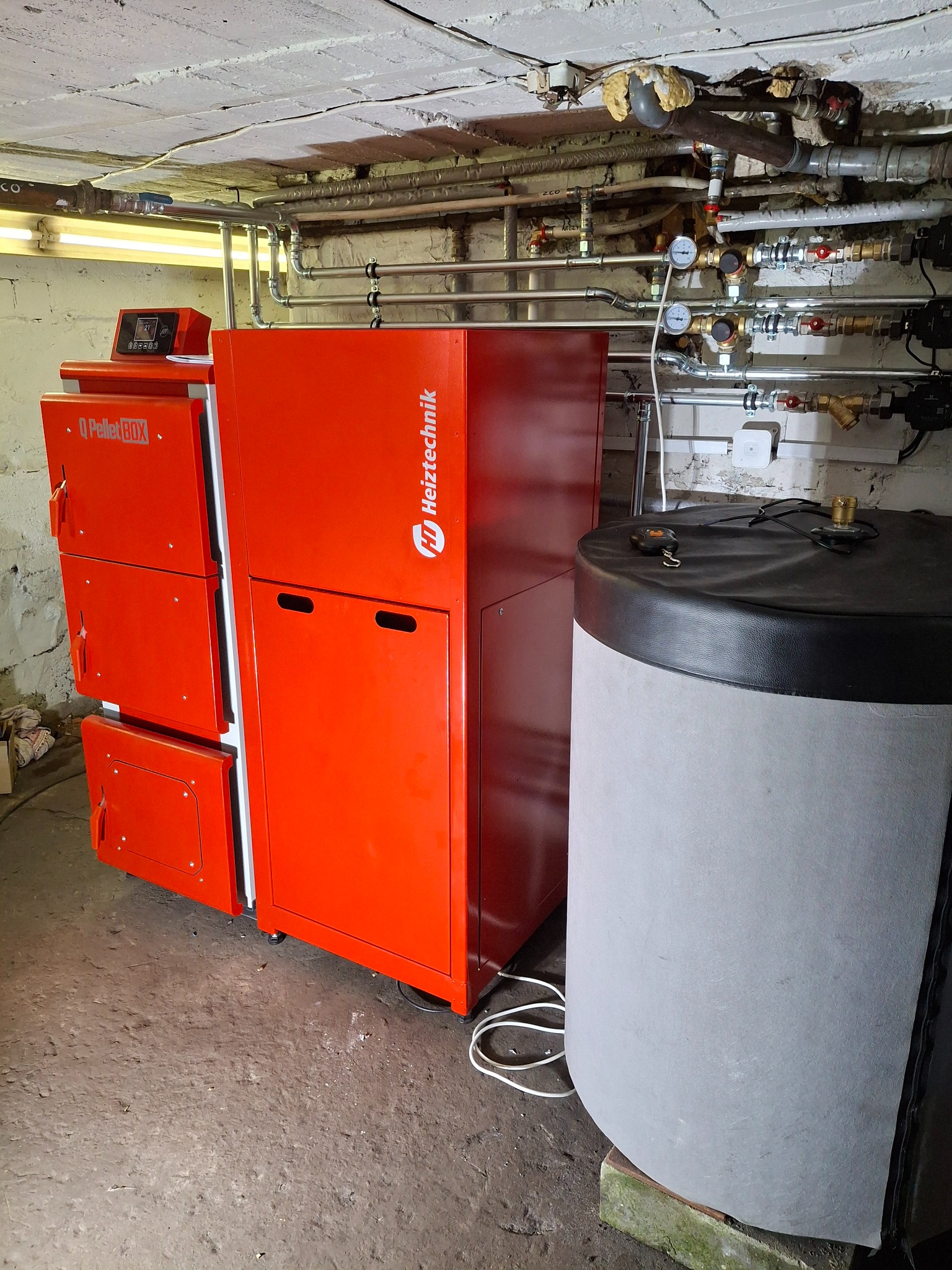 Pomarańczowy piec Heiztechnik Q Pellet BOX z zasobnikiem na wodę w kotłowni. Widoczne rury instalacji grzewczej pod sufitem.