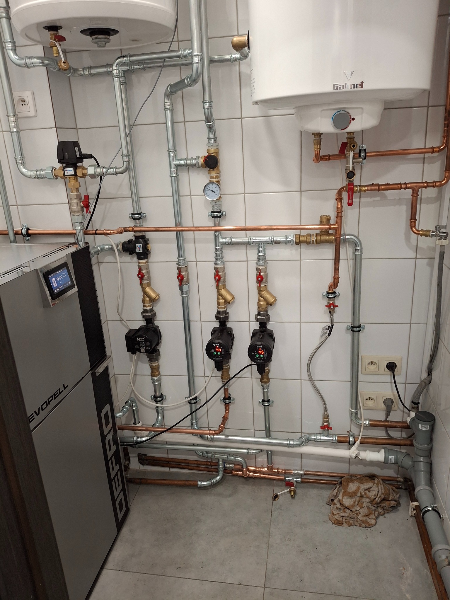 Złożona instalacja grzewcza z kotłem Defro Evopell, zbiornikiem Galmet i licznymi rurami miedzianymi i stalowymi na tle białych płytek. Widoczne pompy obiegowe i zawory.