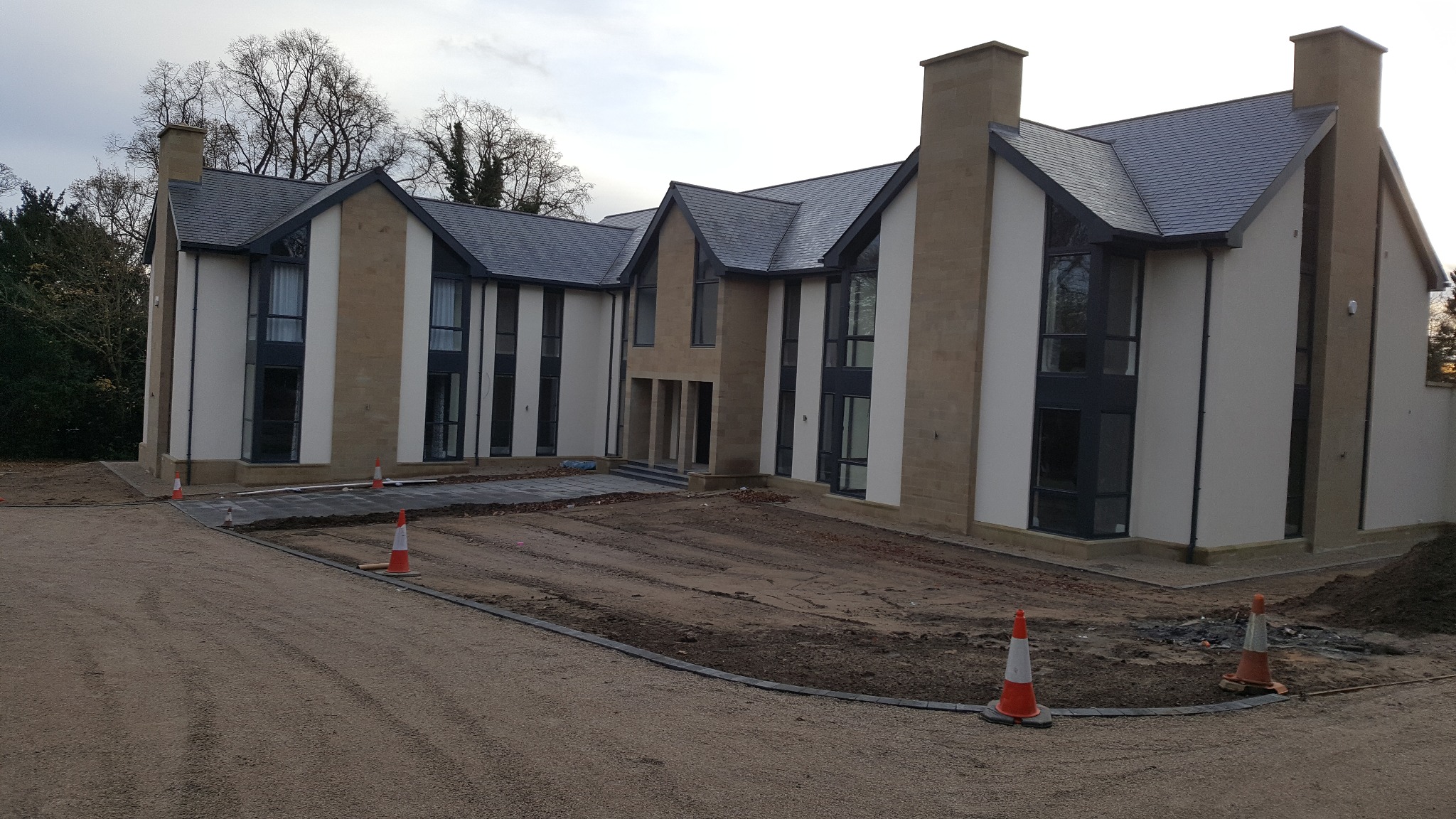 Nowoczesny, dwukondygnacyjny dom z płaskim dachem pokrytym szarą dachówką, z elewacją w kolorze białym i beżowym, duże okna z ciemnymi ramami, teren wokół domu w trakcie prac wykończeniowych...