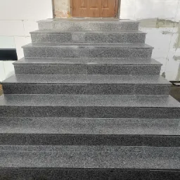 Granitowe schody wejściowe do budynku z drewnianymi drzwiami, ściany z białego bloczka betonowego, widoczne ślady prac budowlanych.