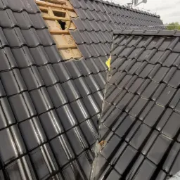 Częściowo zdjęte czarne dachówki na dachu domu, odsłonięta drewniana konstrukcja, widoczna antena satelitarna.