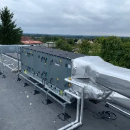 Szara centrala wentylacyjna na dachu budynku z widocznymi przewodami izolowanymi folią aluminiową, tło stanowią drzewa i zabudowania mieszkalne.