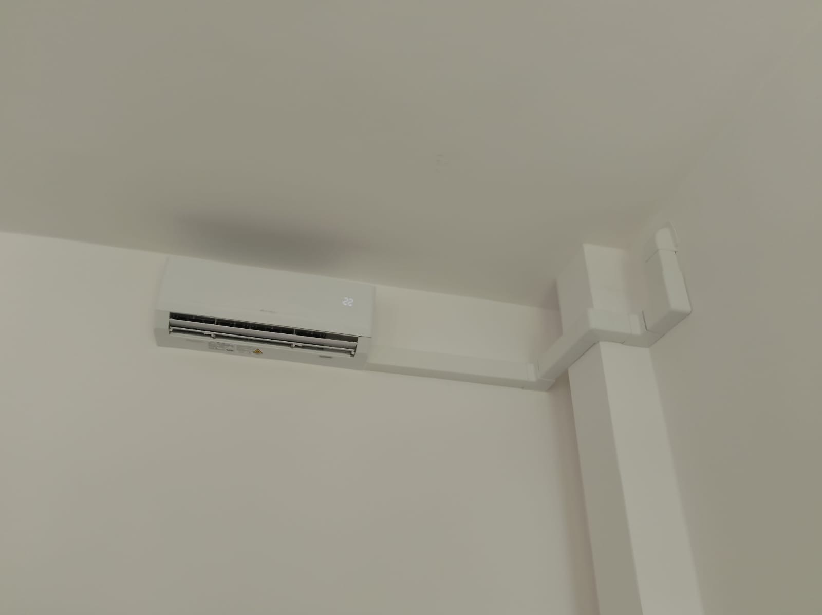Biały klimatyzator ścienny z wyświetlaczem temperatury 22 stopnie Celsjusza, zamontowany na białej ścianie w rogu pomieszczenia, z widocznymi białymi listwami maskującymi instalację.