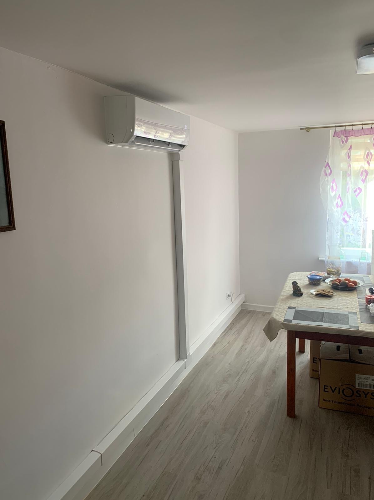 Biały klimatyzator ścienny zamontowany w pokoju mieszkalnym, z widocznymi maskownicami instalacji elektrycznej i chłodniczej, oraz stołem z przekąskami w tle.