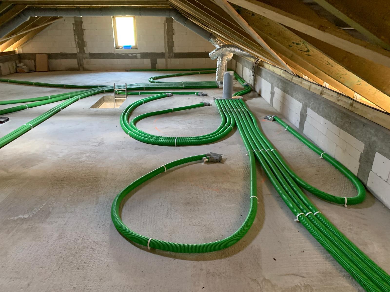Zielone, elastyczne rury wentylacyjne rozprowadzone na poddaszu domu w trakcie instalacji systemu, widoczne drewniane krokwie dachu i okno z naturalnym światłem.