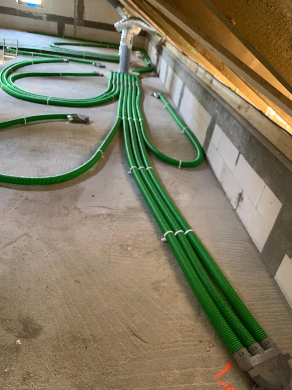 Zielone, karbowane rury wentylacyjne rozprowadzone po podłodze poddasza, połączone w wiązki za pomocą białych opasek zaciskowych, z widocznym systemem mocowania do centralnego przewodu wentylacyjnego.