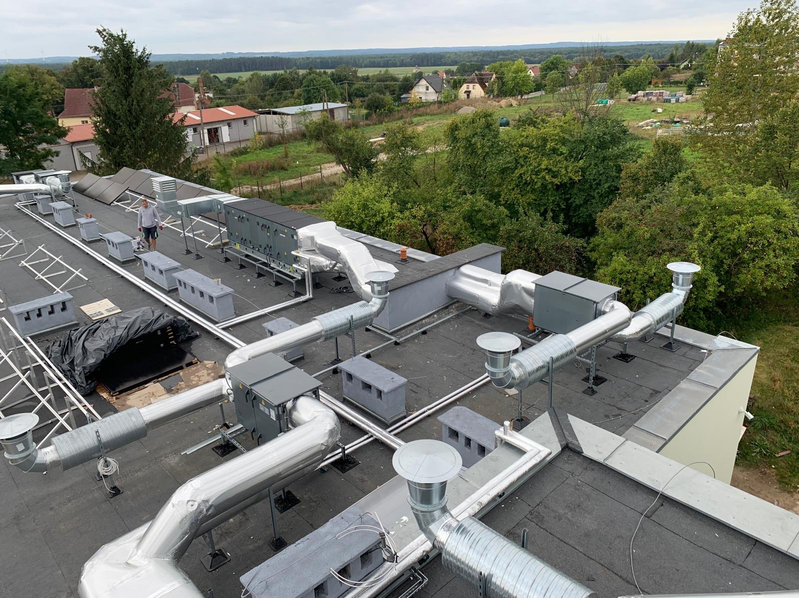 Instalacja wentylacyjna z srebrnymi rurami i szarymi skrzynkami na płaskim dachu budynku, widok z góry na tle krajobrazu wiejskiego i pochmurnego nieba, pracownik w oddali.