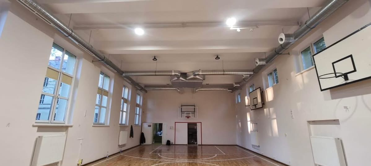 Sala gimnastyczna z widoczną instalacją wentylacyjną pod sufitem. Metalowe rury wentylacyjne biegną wzdłuż sufitu, z nawiewnikami. Widoczne okna i kosze do koszykówki.