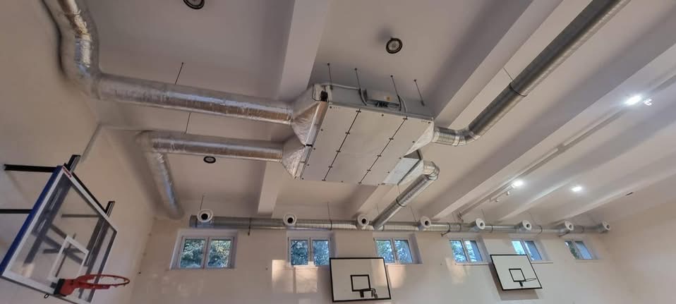 Instalacja rekuperacji w sali gimnastycznej: srebrne rury wentylacyjne pod sufitem, centrala rekuperacyjna, okna i kosze do koszykówki w tle.