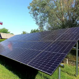 Instalacja paneli słonecznych na trawie, w tle budynek z polską flagą, błękitne niebo i drzewa.