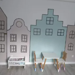 Pokój dziecięcy z ręcznie malowaną ścianą przedstawiającą budynki w stylu holenderskim, biały stolik i krzesełka z kolorowymi nogami, oraz biały regał na zabawki.