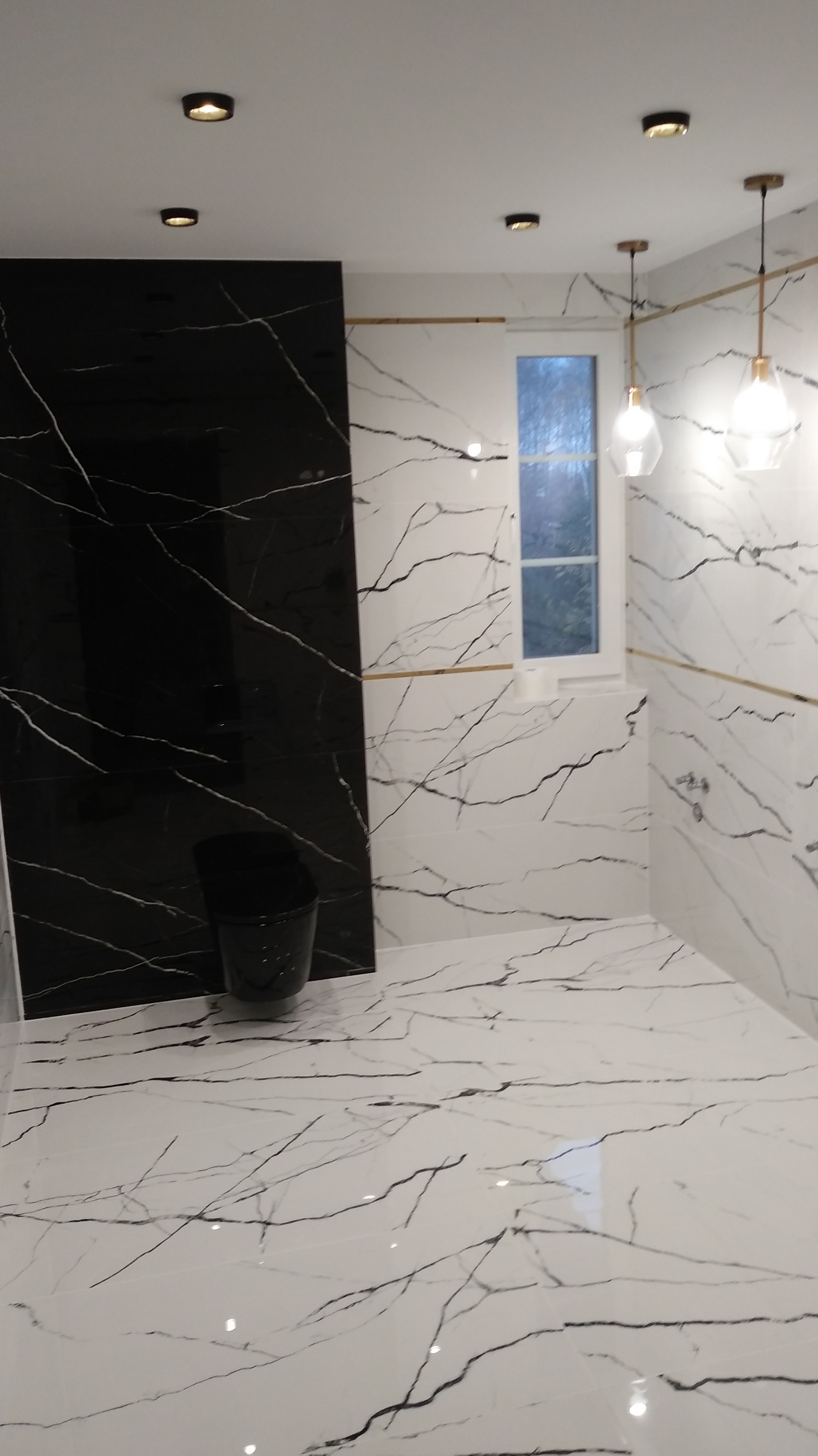 Wykończona łazienka z czarno-białymi płytkami imitującymi marmur, podwieszana toaleta w kolorze czarnym, oświetlenie sufitowe punktowe i wiszące lampy z przezroczystymi kloszami, okno z widokiem...