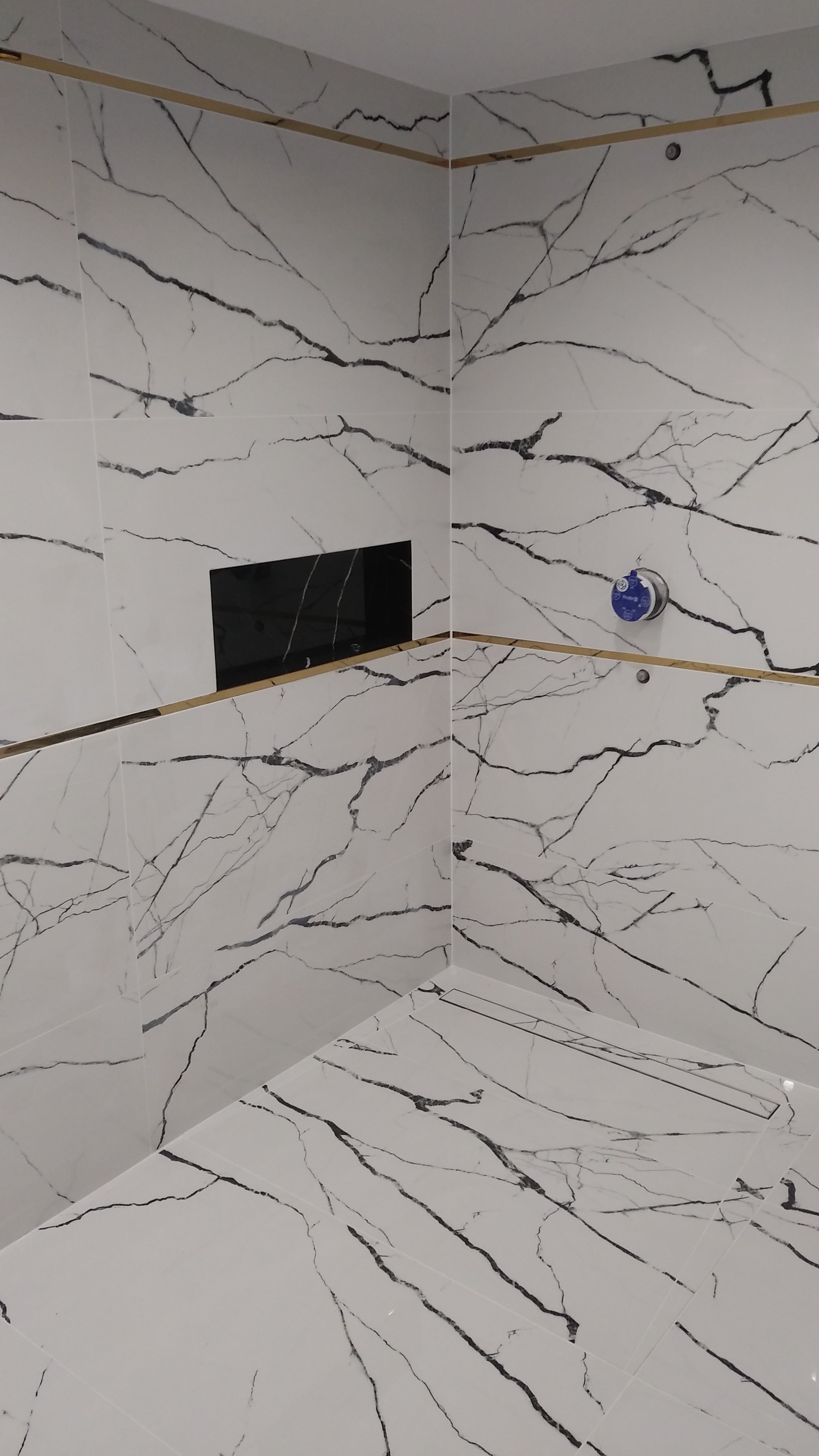 Wykończona łazienka z białymi płytkami imitującymi marmur, złotymi listwami dekoracyjnymi i czarną wnęką na kosmetyki.