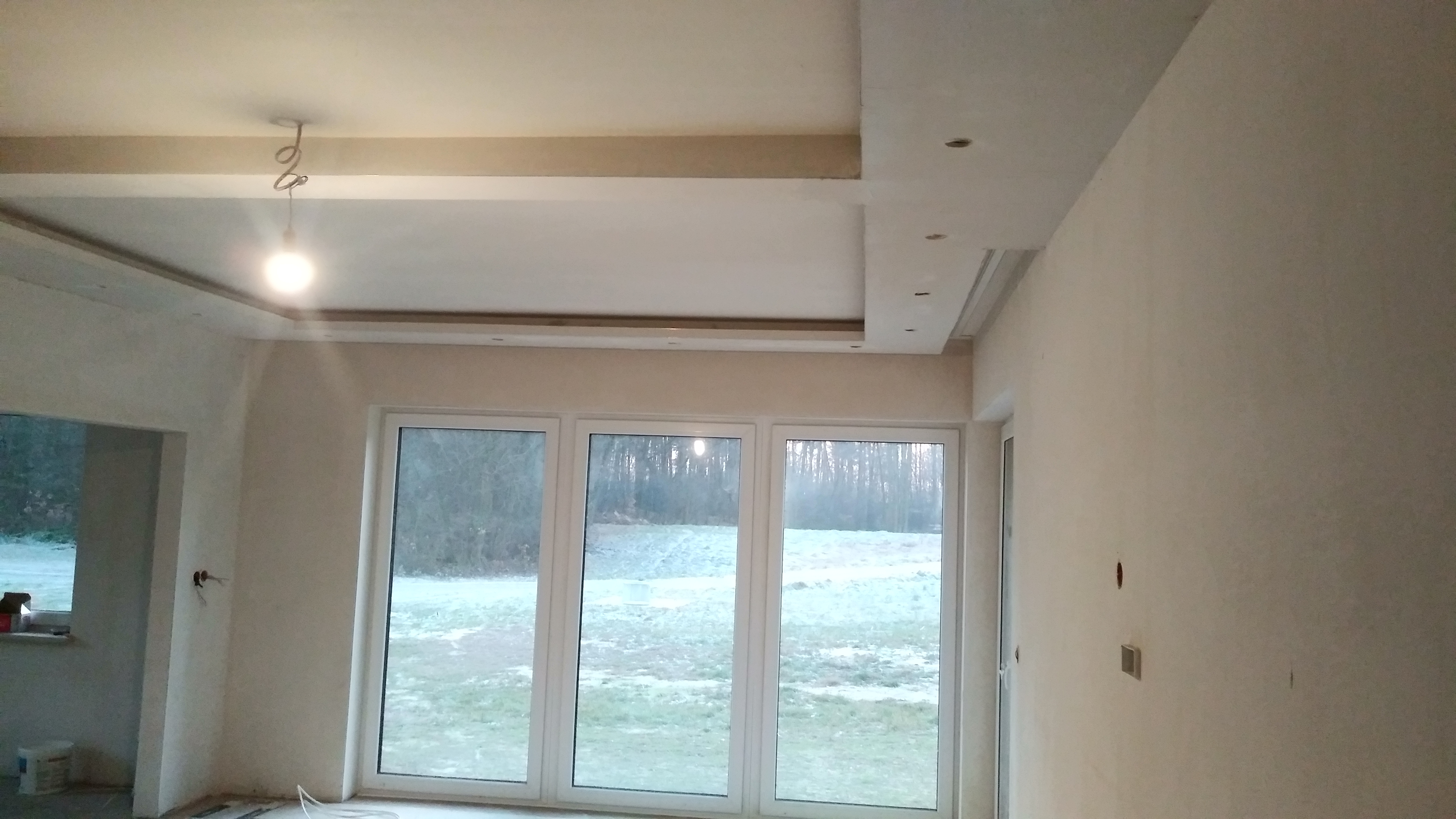 Wnętrze w trakcie prac wykończeniowych: sufit podwieszany z oświetleniem punktowym, duże okna z widokiem na zewnątrz, widoczne niedokończone instalacje elektryczne w ścianach.