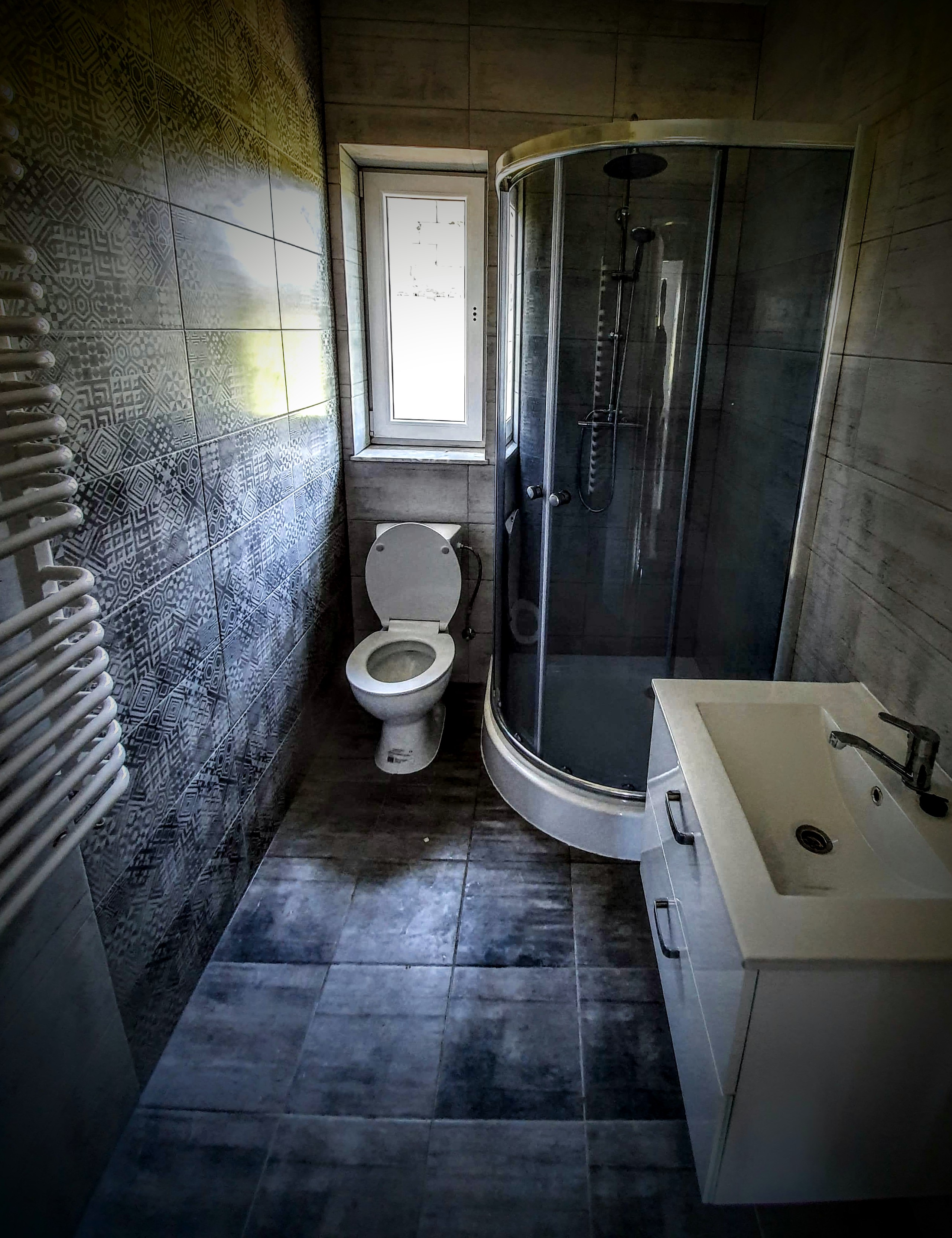 Wnętrze łazienki z kabiną prysznicową narożną, toaletą i umywalką z szafką w szaro-białej kolorystyce, z dekoracyjnymi płytkami na ścianie.