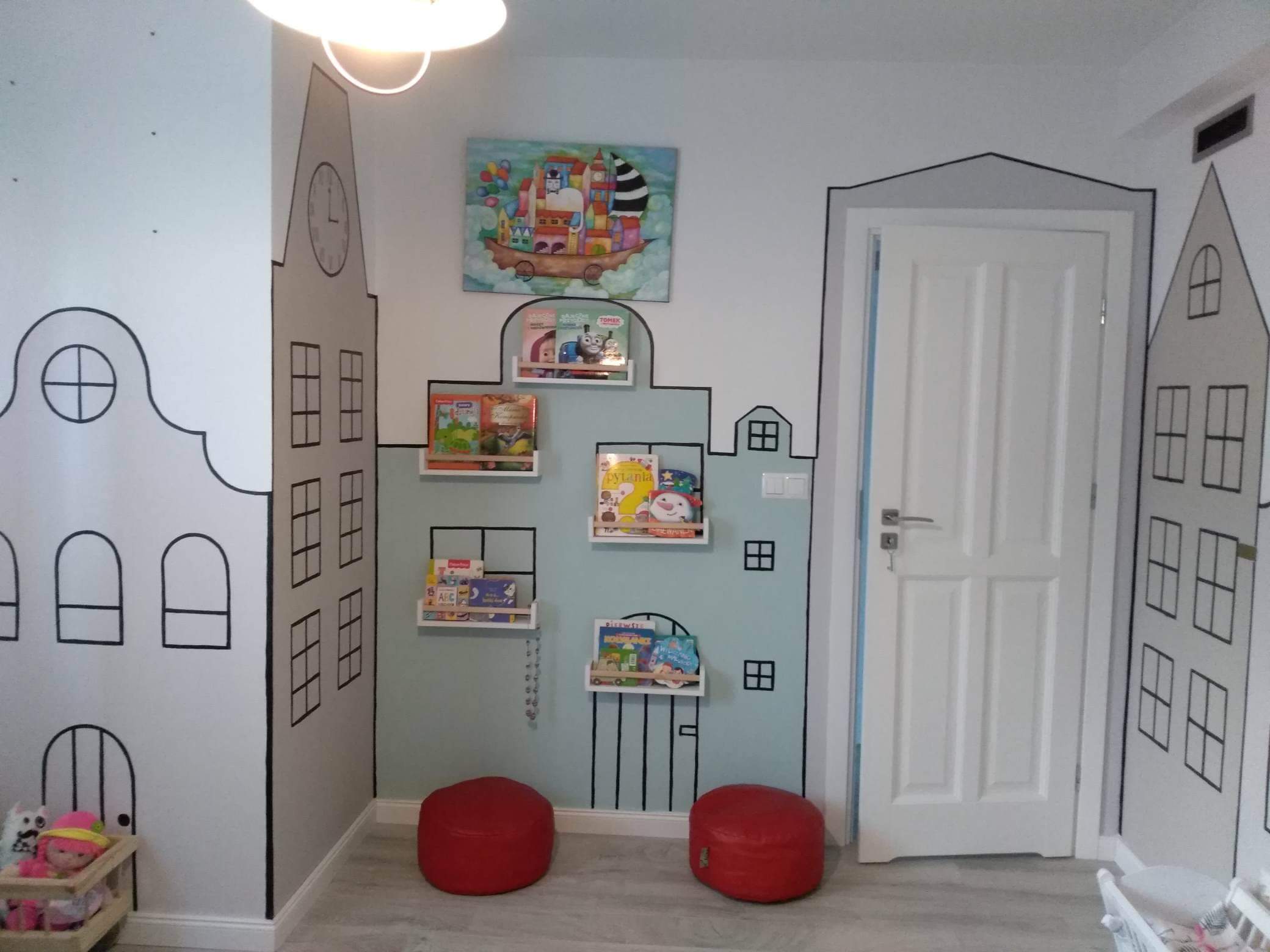 Pokój dziecięcy z ręcznie malowanymi motywami miejskimi na ścianach, półkami na książki w kształcie okien i dwoma czerwonymi pufami na podłodze.