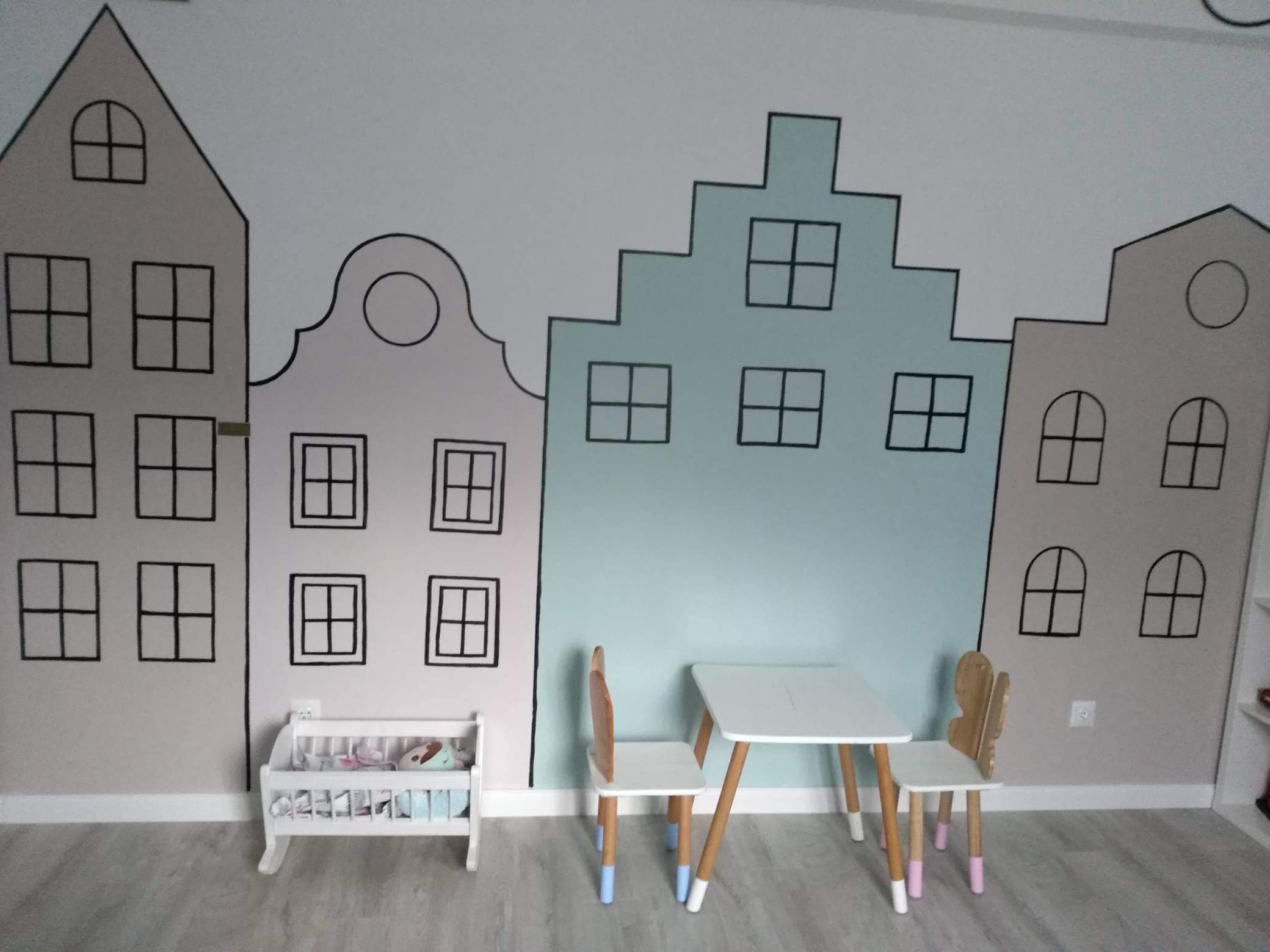 Pokój dziecięcy z ręcznie malowaną ścianą przedstawiającą budynki w stylu holenderskim, biały stolik i krzesełka z kolorowymi nogami, oraz biały regał na zabawki.