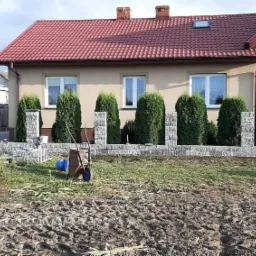 Kamienny murek ogrodzeniowy z elementami słupków w Gdańsku, z tłem domu z czerwoną dachówką i tujami, widoczny wózek budowlany.