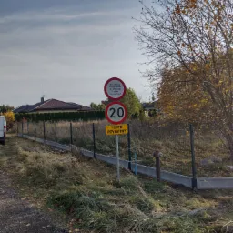 Metalowa siatka ogrodzeniowa z betonową podmurówką oddzielająca teren prywatny, widoczne znaki drogowe i jesienne drzewo.