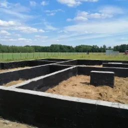 Zabezpieczone hydroizolacją fundamenty z bloczków betonowych, wypełnione piaskiem, na tle zieleni i błękitnego nieba z chmurami.