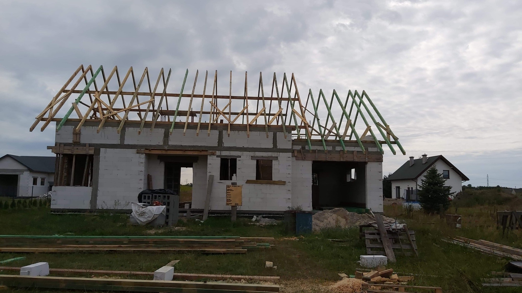 Niedokończony budynek z białych bloczków z drewnianą konstrukcją dachu, częściowo zaimpregnowaną na zielono, na tle pochmurnego nieba i innych domów w oddali.
