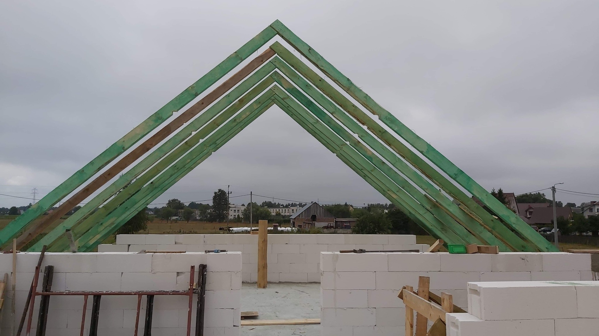 Wznoszenie konstrukcji dachu z impregnowanych na zielono belek na budynku z białych bloczków, widok na okolicę w pochmurny dzień.
